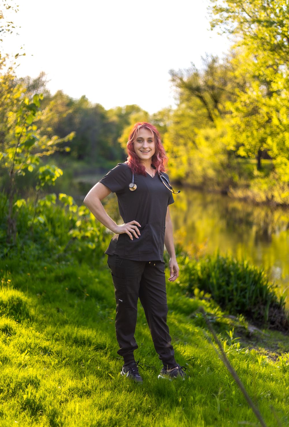 Healthcare worker in black scrubs standing on grassy riverbank during golden hour.