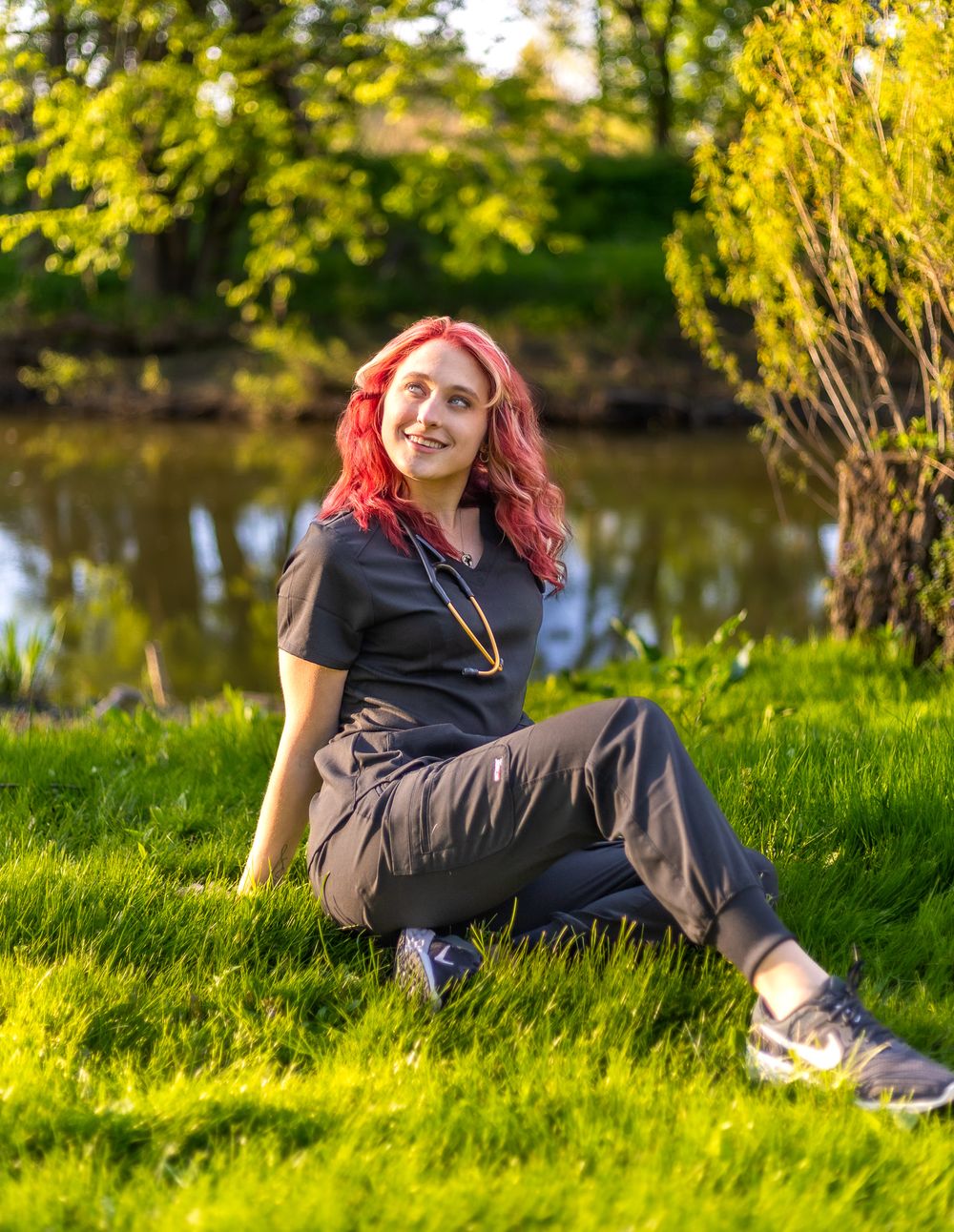 Person in casual attire with bright hair relaxing on grass by a tranquil pond on a sunny day.