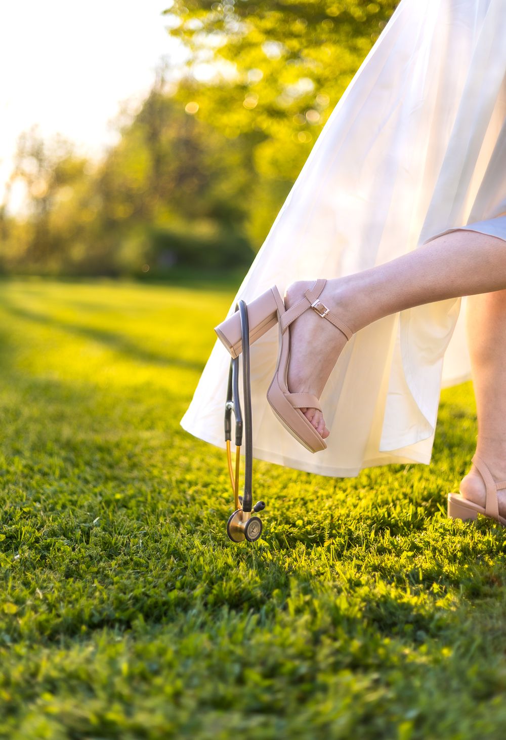 Close-up of elegant beige heels on grass during golden hour with white dress fabric flowing.