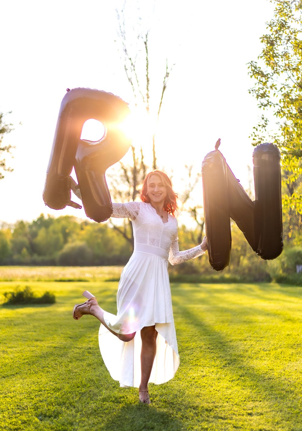 Person in white dress jumping with large letter balloons spelling 'RN' during sunset in a grassy field.