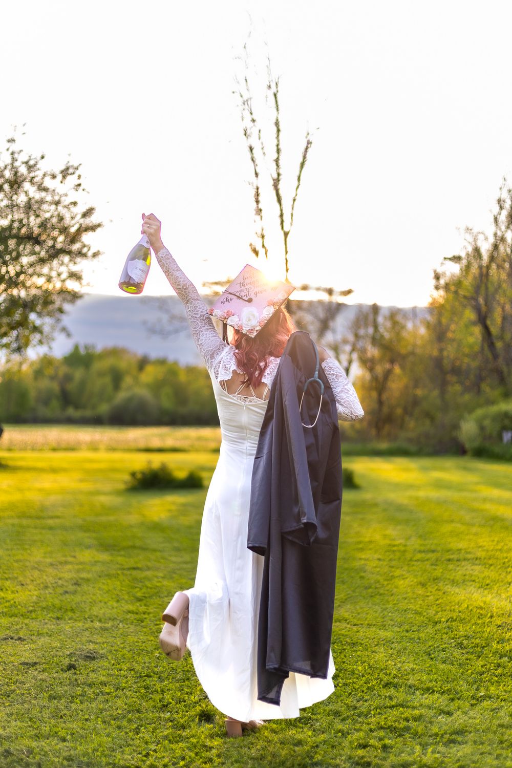 Figure in white dress holding up graduation cap and gown against sunset in meadow.