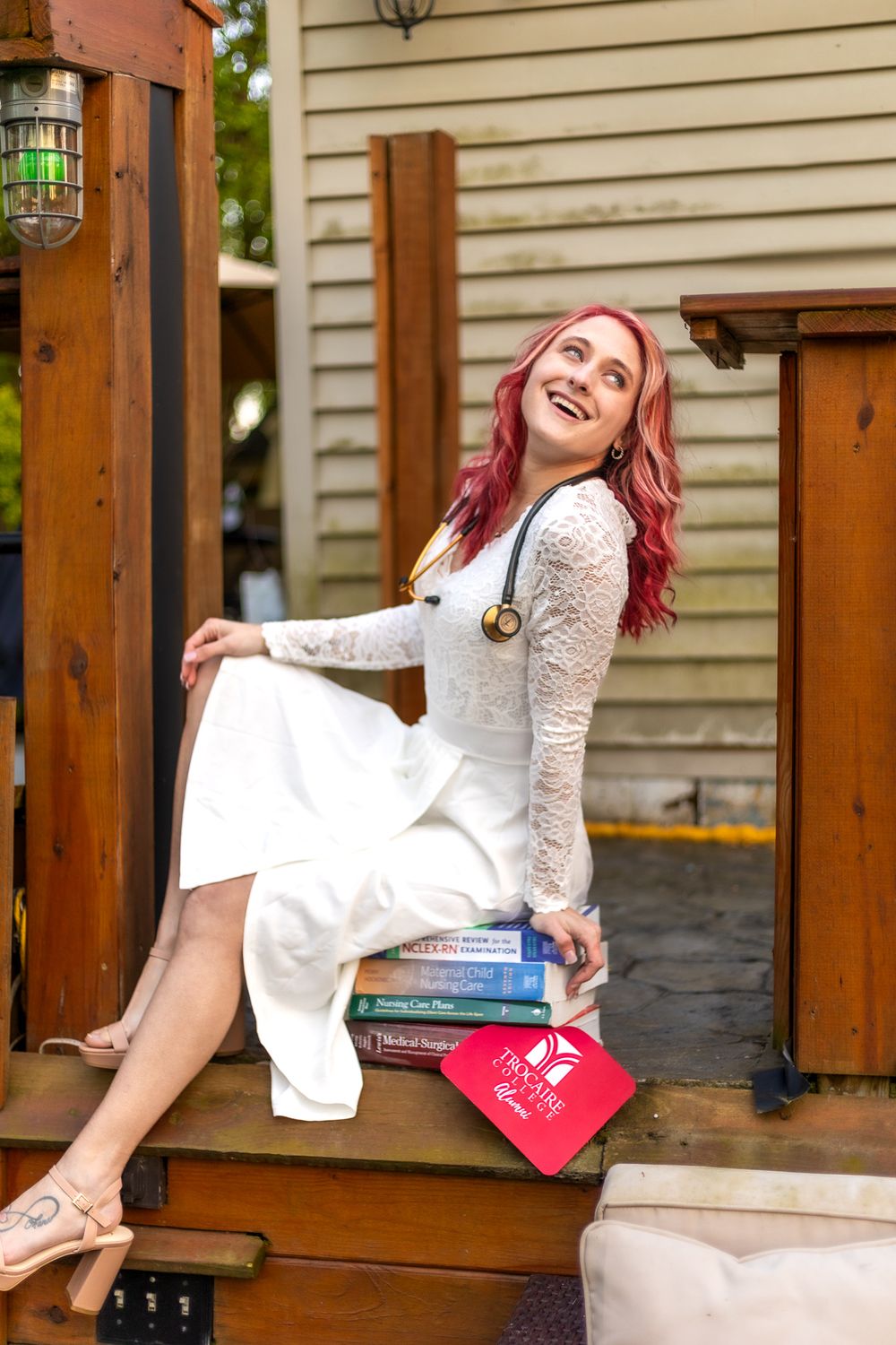 Person in white dress sitting on wooden porch steps with books and bright hair.