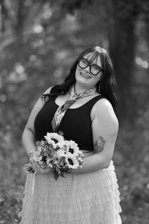 Black and white portrait of someone in a sleeveless dress with ruffled layers holding a floral bouquet in a forest setting.