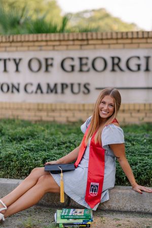 A graduate in red and grey attire poses near a brick wall and greenery at a university campus.