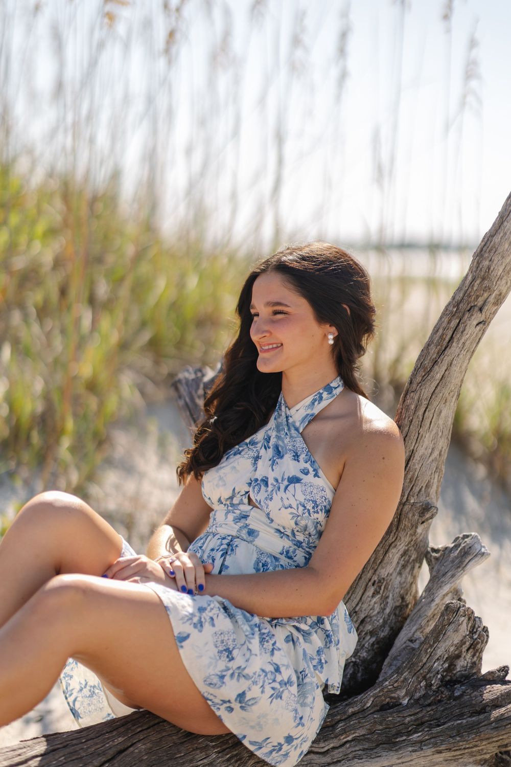 Young woman in blue and white floral halter dress sits on driftwood at beach during golden hour.