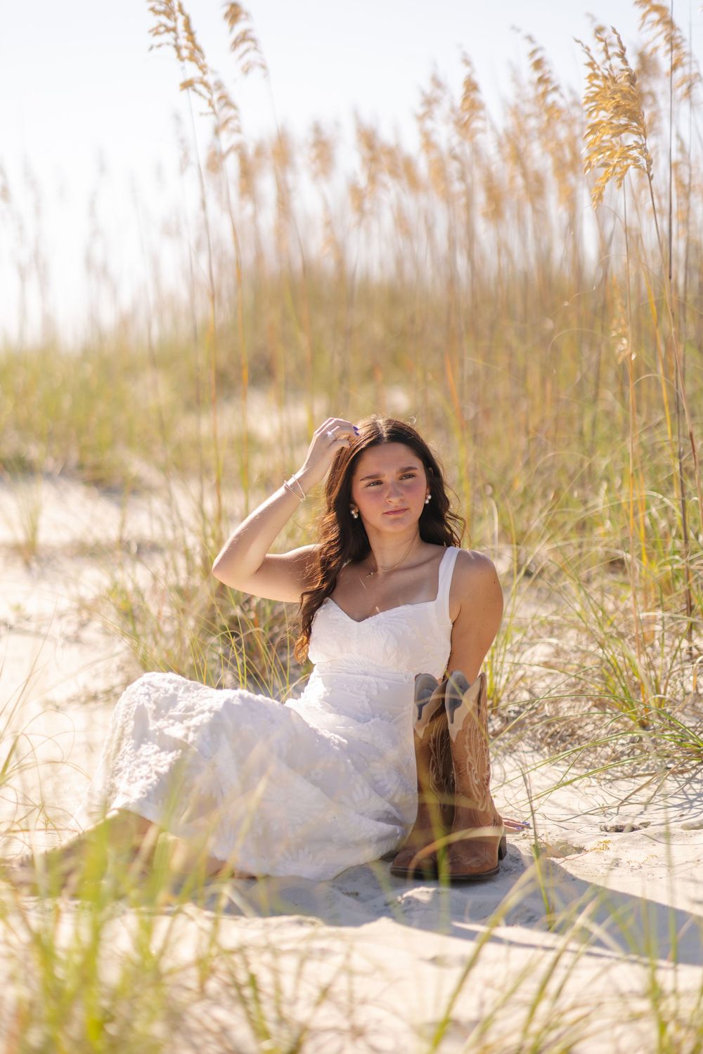 Person in white sundress sitting among beach grass and sand dunes in warm sunlit coastal setting.
