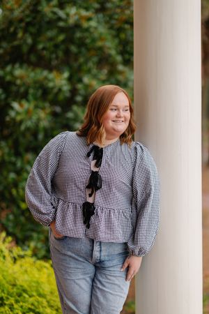 A person wearing a black and white gingham top and jeans poses playfully against a white column outdoors.