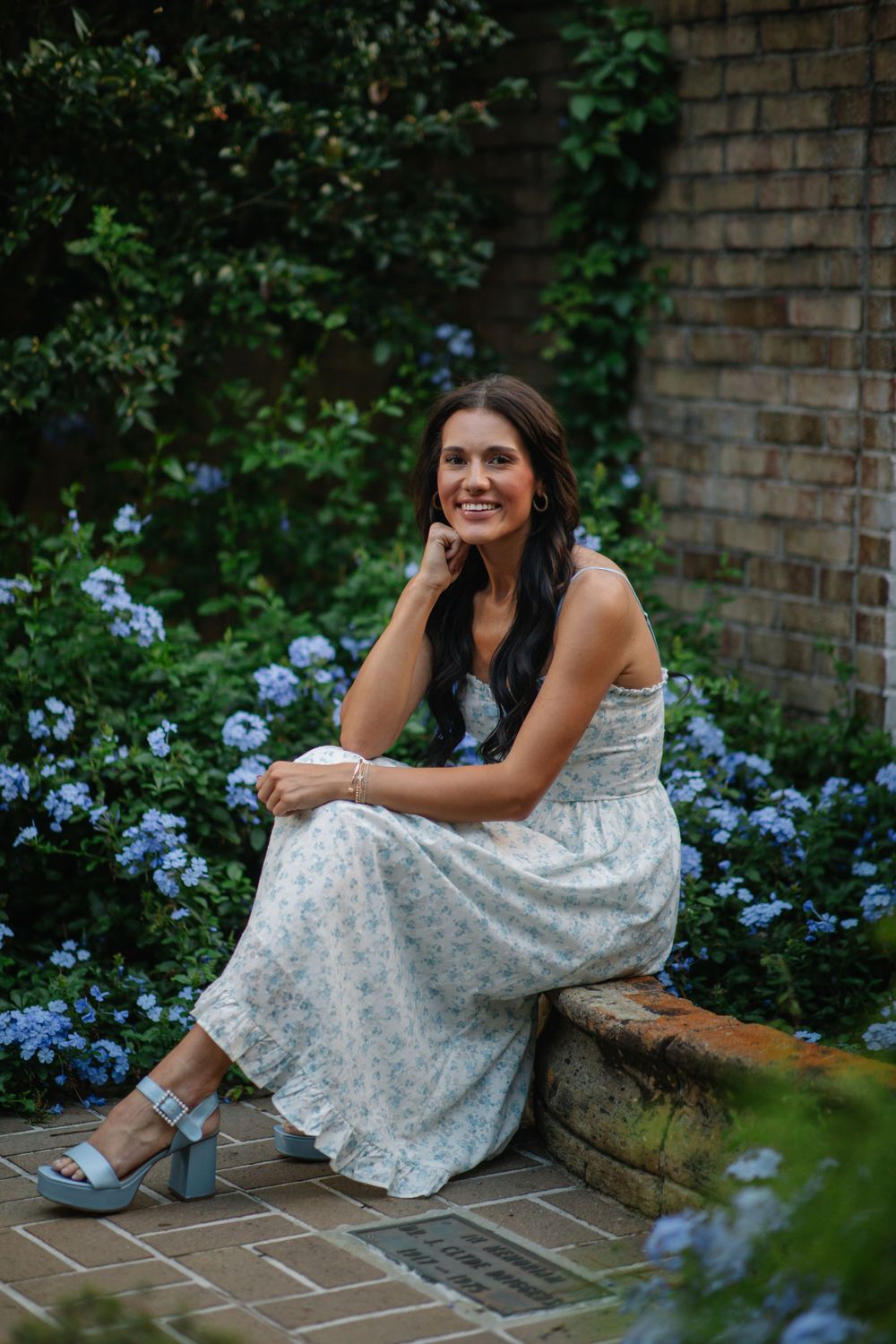 A figure in a floral white dress sits on a garden bench surrounded by blooming blue hydrangeas and brick walls.