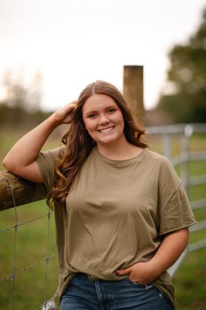 A person in a tan t-shirt and jeans poses casually by a wooden fence in an outdoor rural setting at sunset.