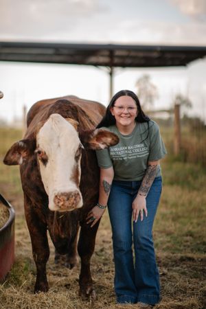 A person in a green t-shirt and jeans poses with a large brown and white cow in a rural farm setting.