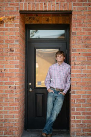 Someone casually leans in a black doorway of a red brick building wearing a plaid shirt and jeans.