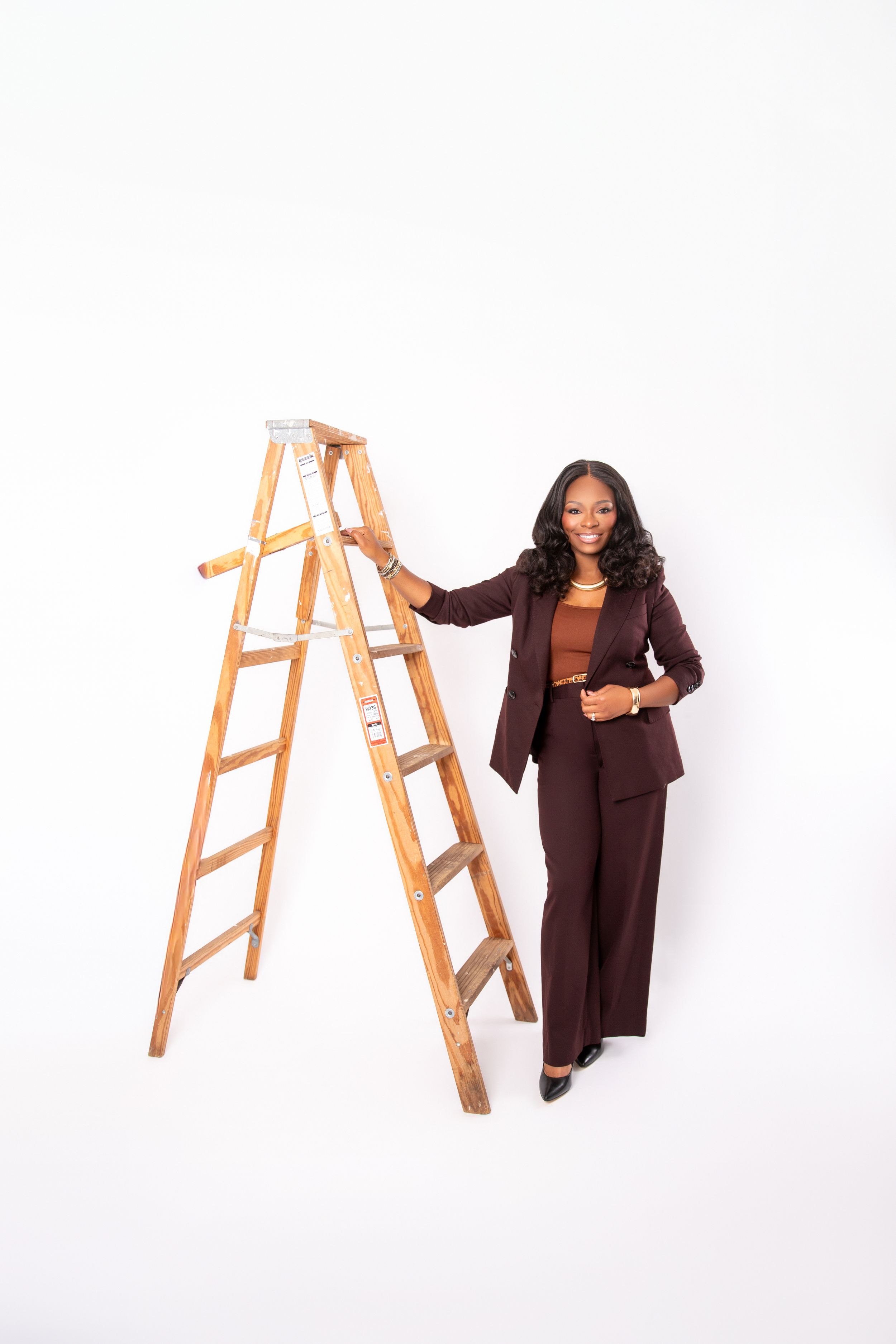 Confident businesswoman in brown suit standing beside wooden ladder against white background.
