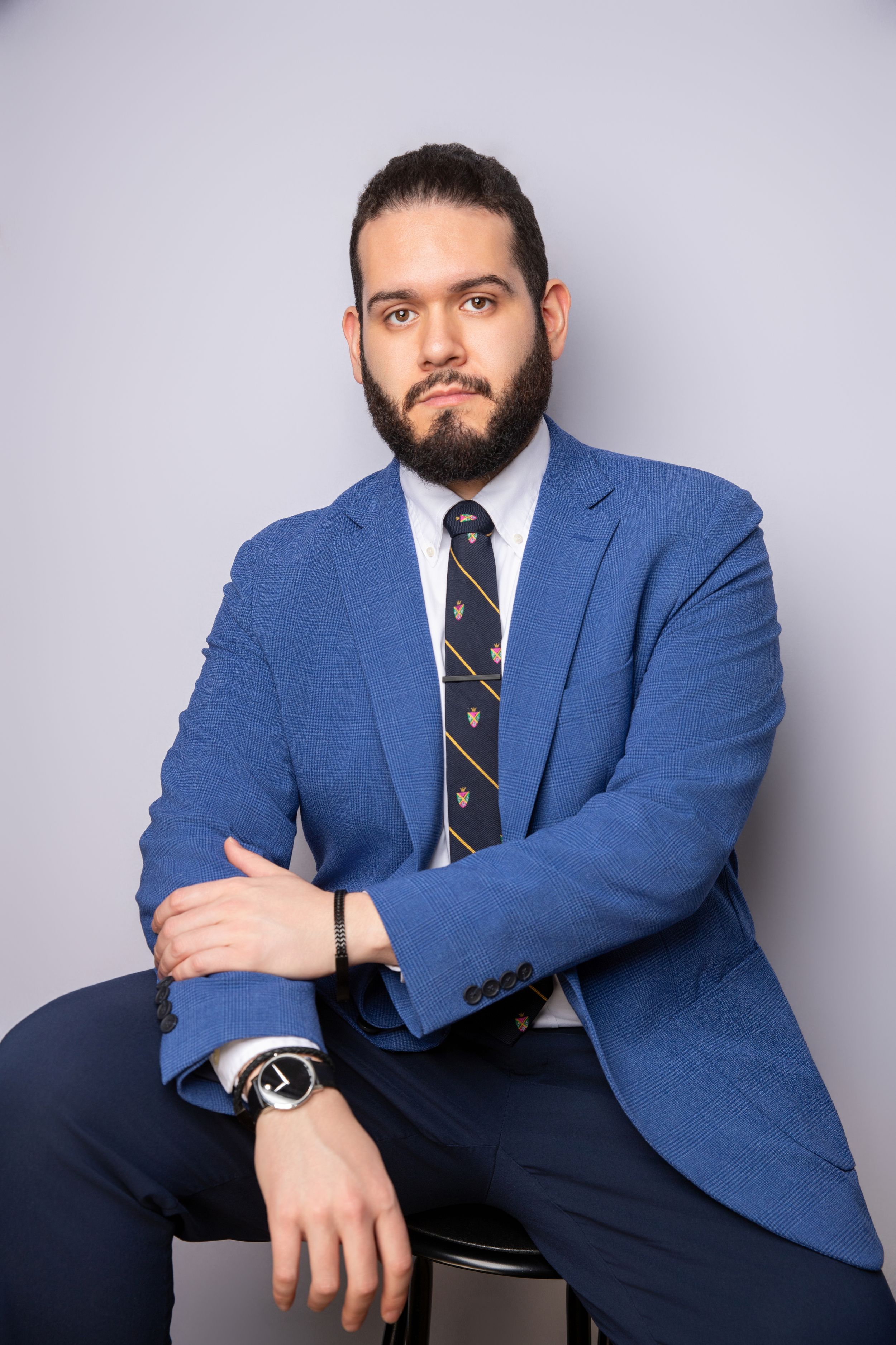 Professional headshot of person in blue suit jacket and striped tie against grey background.