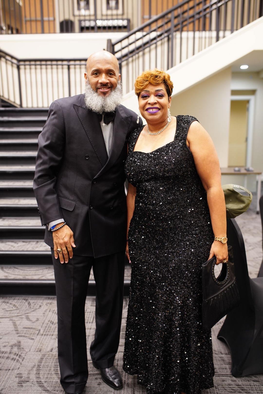 A couple dressed in formal attire, with the gentleman in a tuxedo and the lady wearing a sparkling black evening gown.
