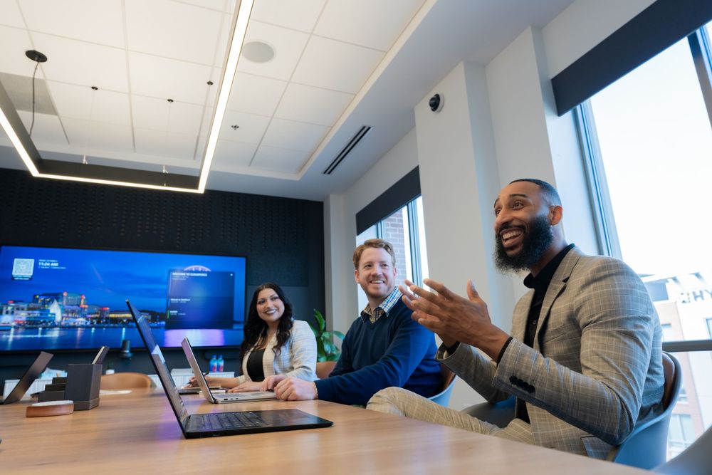 Three professionals laughing and collaborating in a modern conference room with a laptop and large display screen.