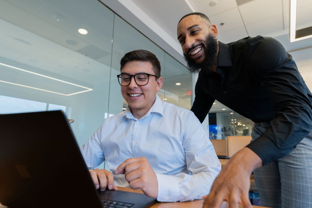 Two businessmen smiling at a laptop in a modern office, collaborating on work together.