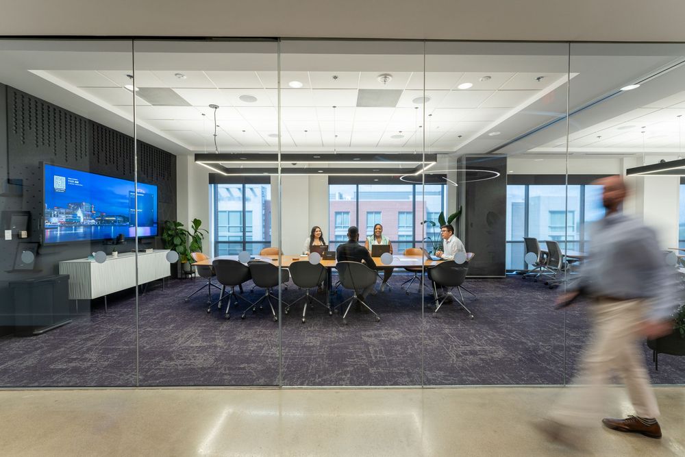 Modern glass-walled conference room with professionals meeting around a oval table, large display screen, and purple carpet.