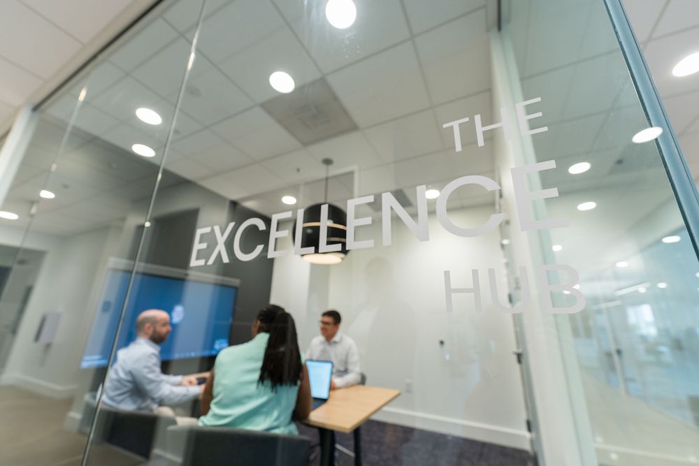 Two professionals meeting in a modern glass-walled office room labeled The Excellence Hub.