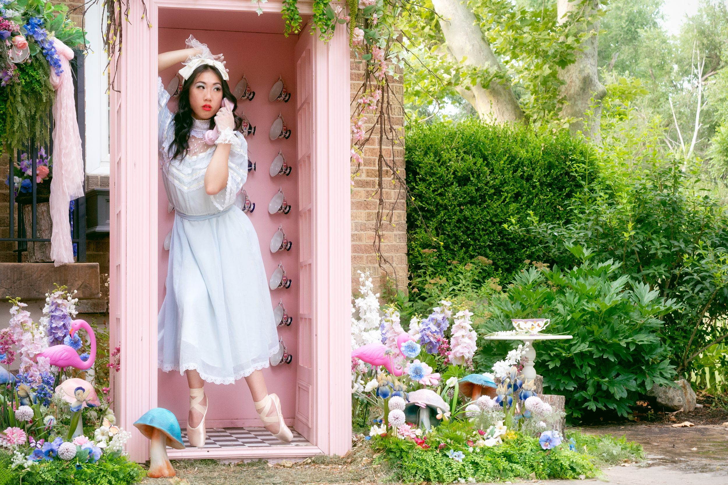 En pointe ballerina poses in pink phone both surrounded by an enchanting garden of flowers and mushrooms with flamingos and tea cups.