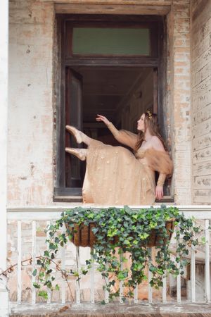 Ballet dancer in golden dress performs graceful pose in window of Race & Religion in New orleans with ivy-covered railing below.