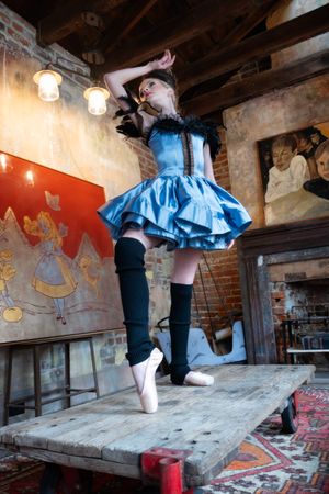 A ballet dancer in a blue dress and black stockings performs en pointe on a wooden platform in a rustic interior setting in New Orleans.