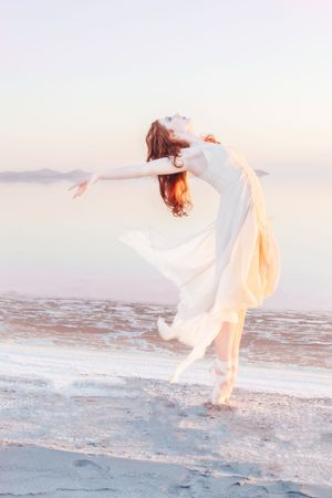 A ballerina in a flowing white dress dances gracefully along the pink water beach of Great Salt Lake at sunset, creating an ethereal scene.