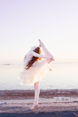 A red haired dancer in a flowing white dress performs graceful movements on a beach at sunset.