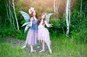 Two sisters in fairy costumes with wings dance together in a sunlit forest clearing in Aspen Grove.