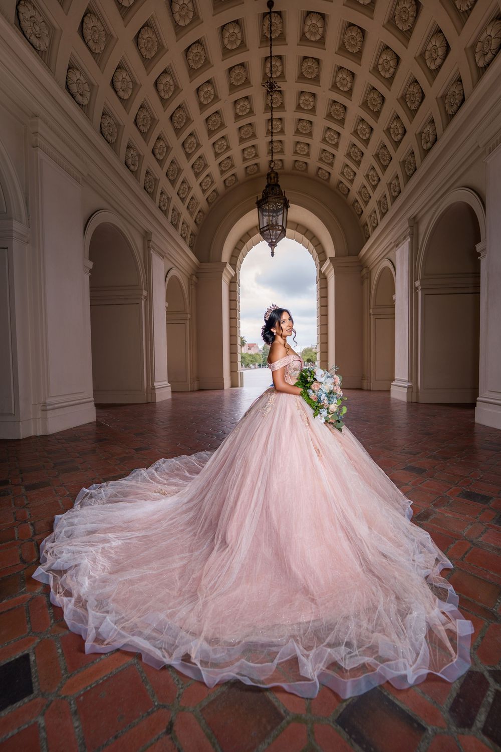 Bride in blush pink ball gown with long train stands in ornate arched corridor holding floral bouquet.