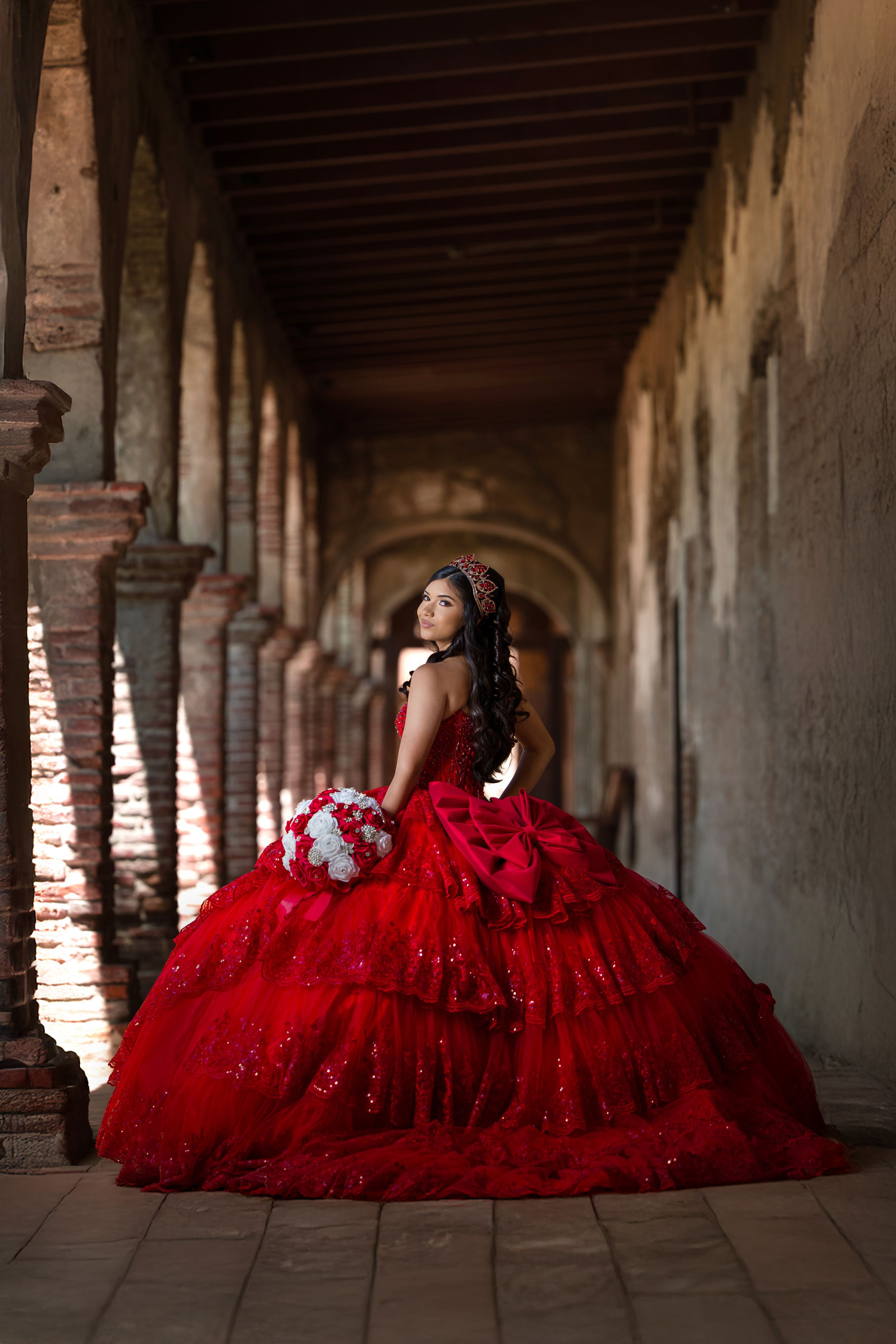 A person in a vibrant red quinceañera ballgown poses elegantly in an arched colonnade hallway with dramatic lighting.