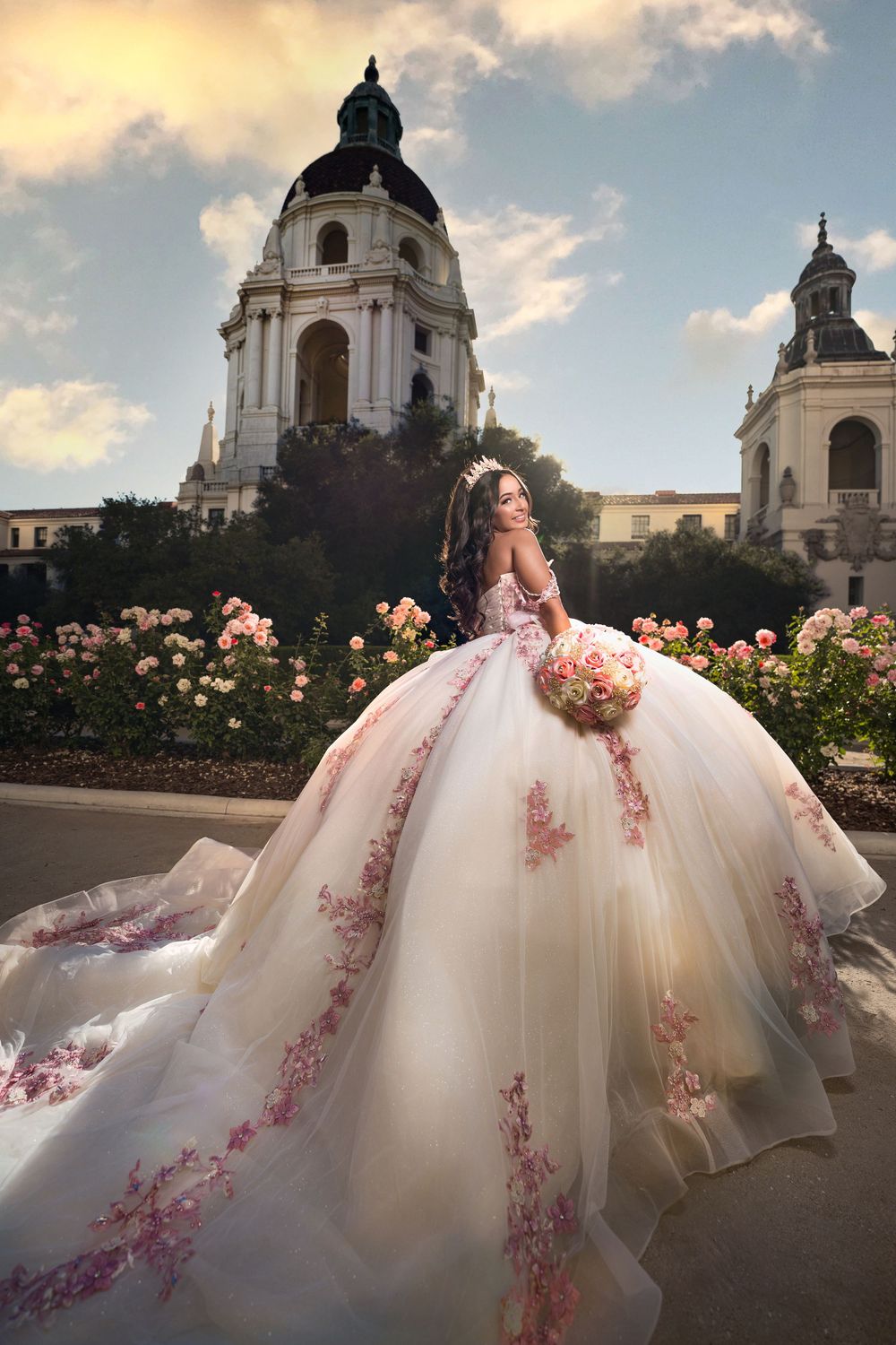 A flowing white ballgown with pink floral embellishments is photographed against a grand architectural backdrop at sunset.