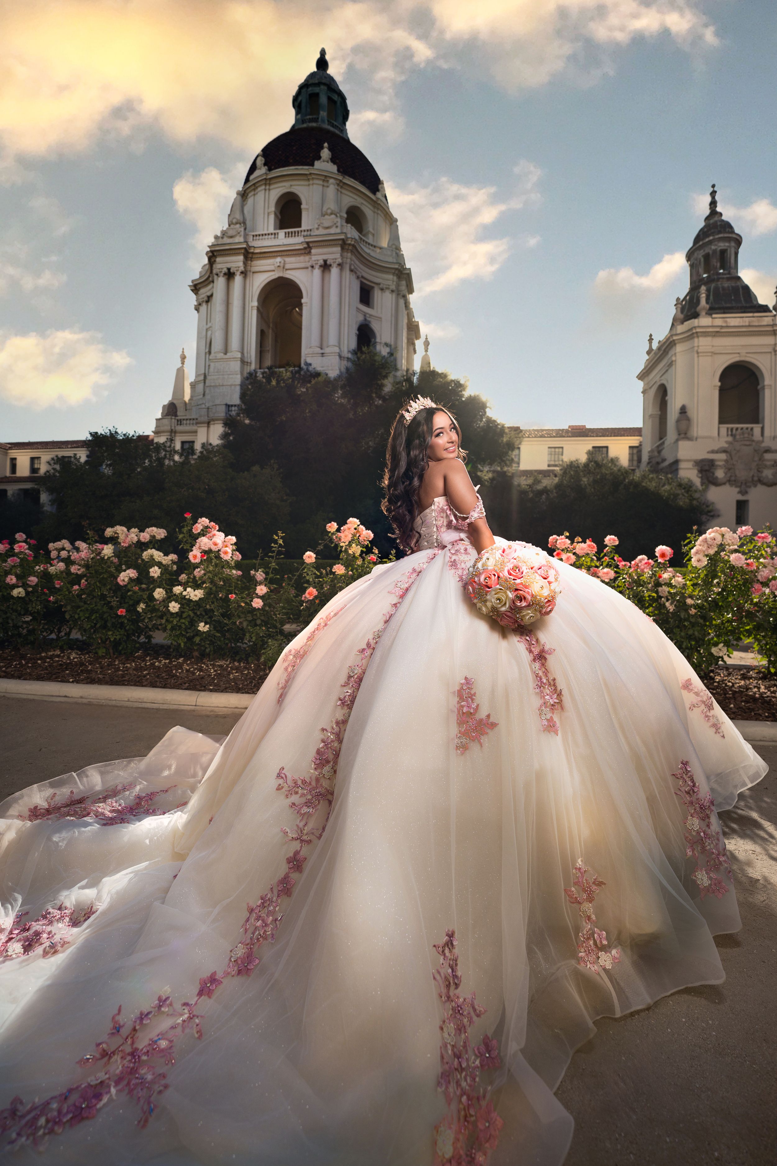 A flowing white ballgown with pink floral embellishments is photographed against a grand architectural backdrop at sunset.