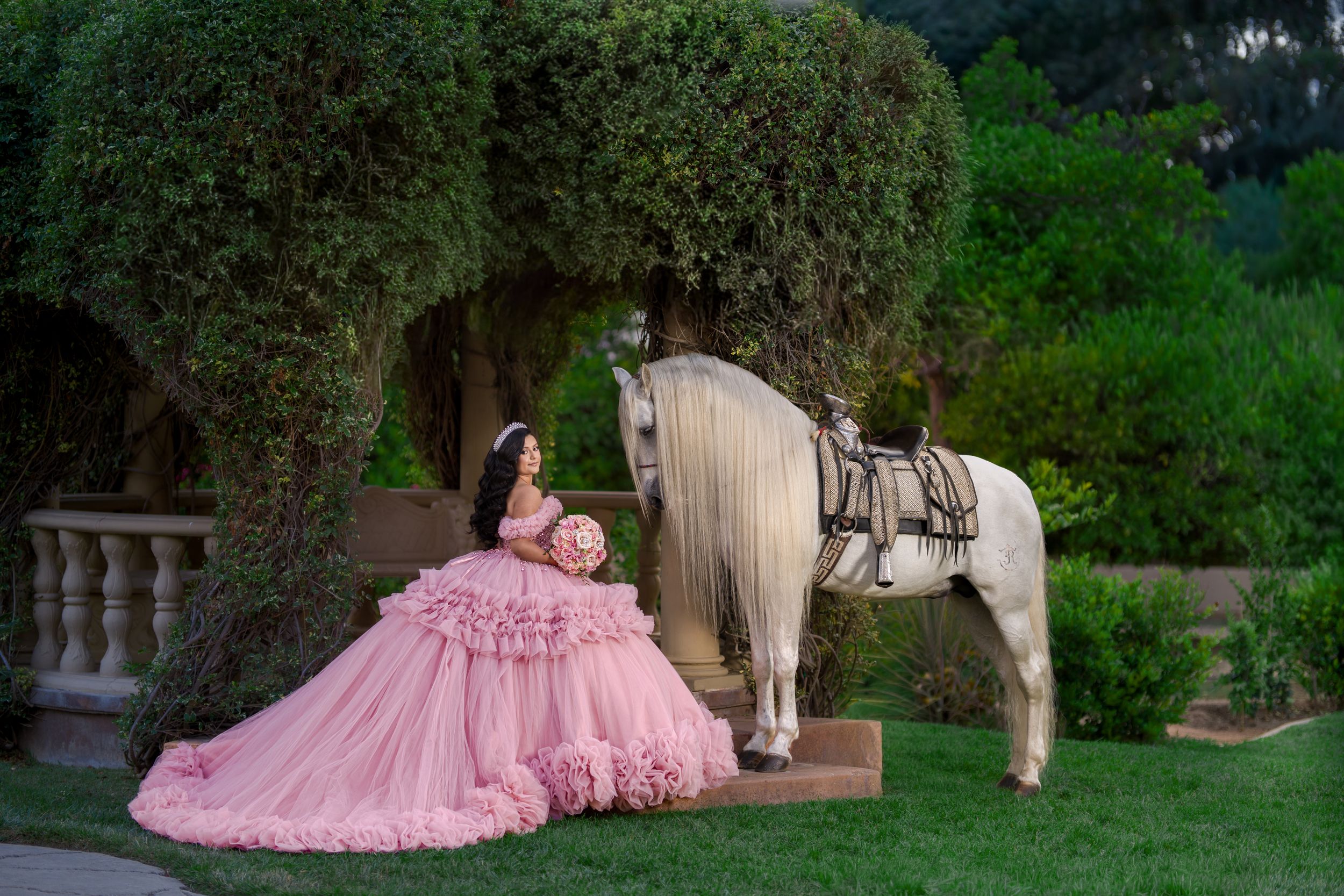 Young woman in pink ball gown poses with white horse in lush garden for quinceañera celebration photo.