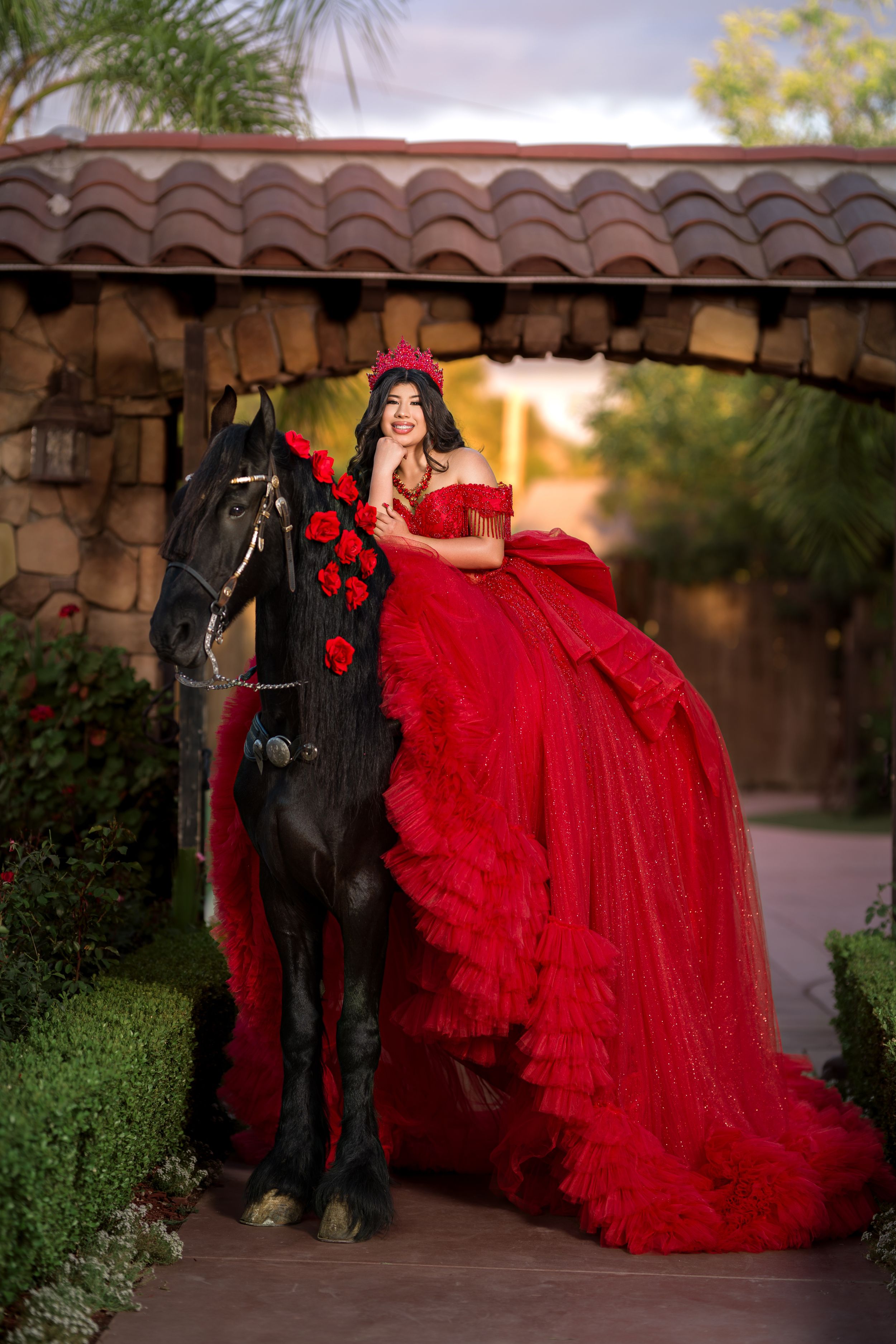 Young woman in red ball gown and crown poses with black horse adorned with roses under stone archway.