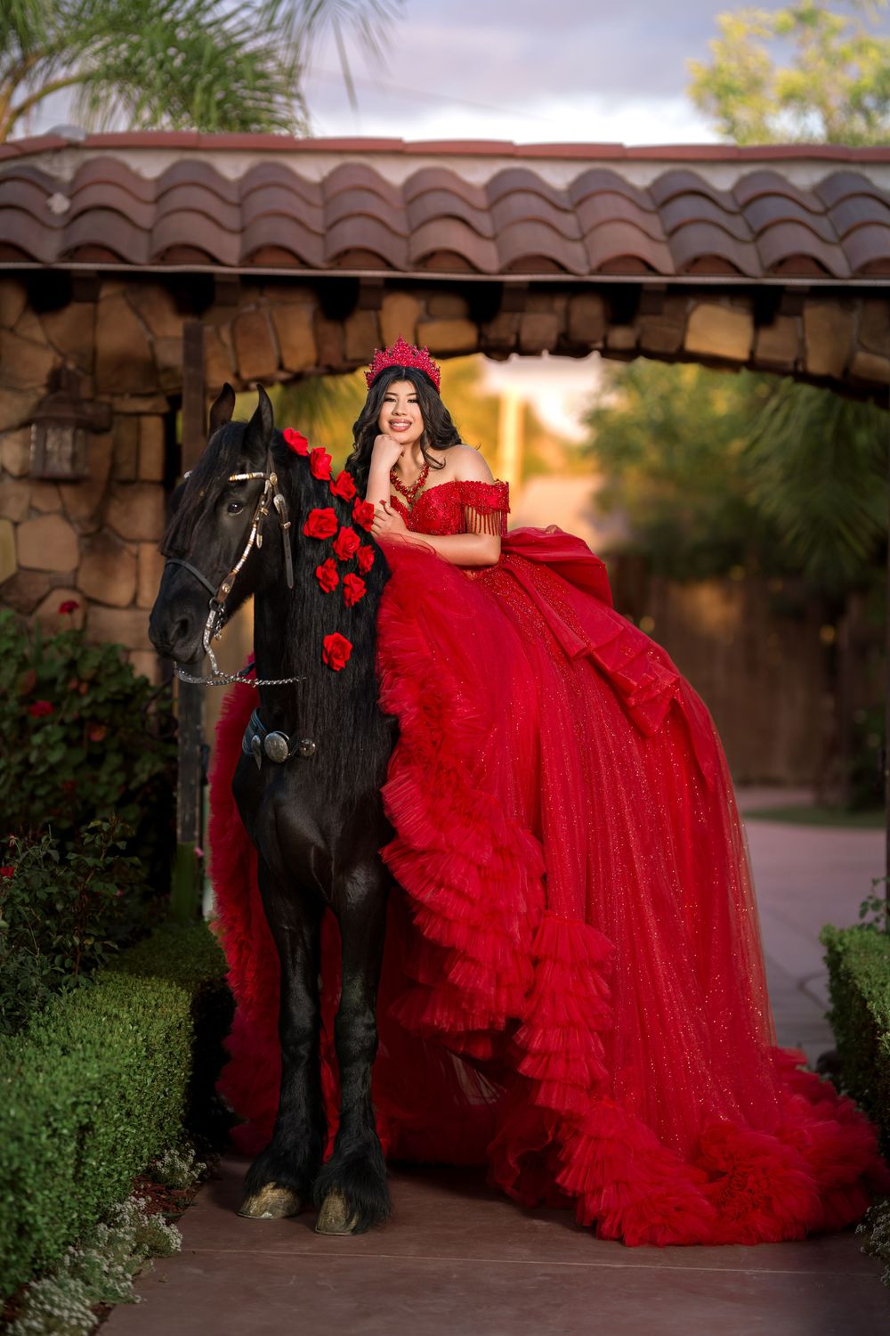 Young woman in red ball gown and crown poses with black horse adorned with roses under stone archway.