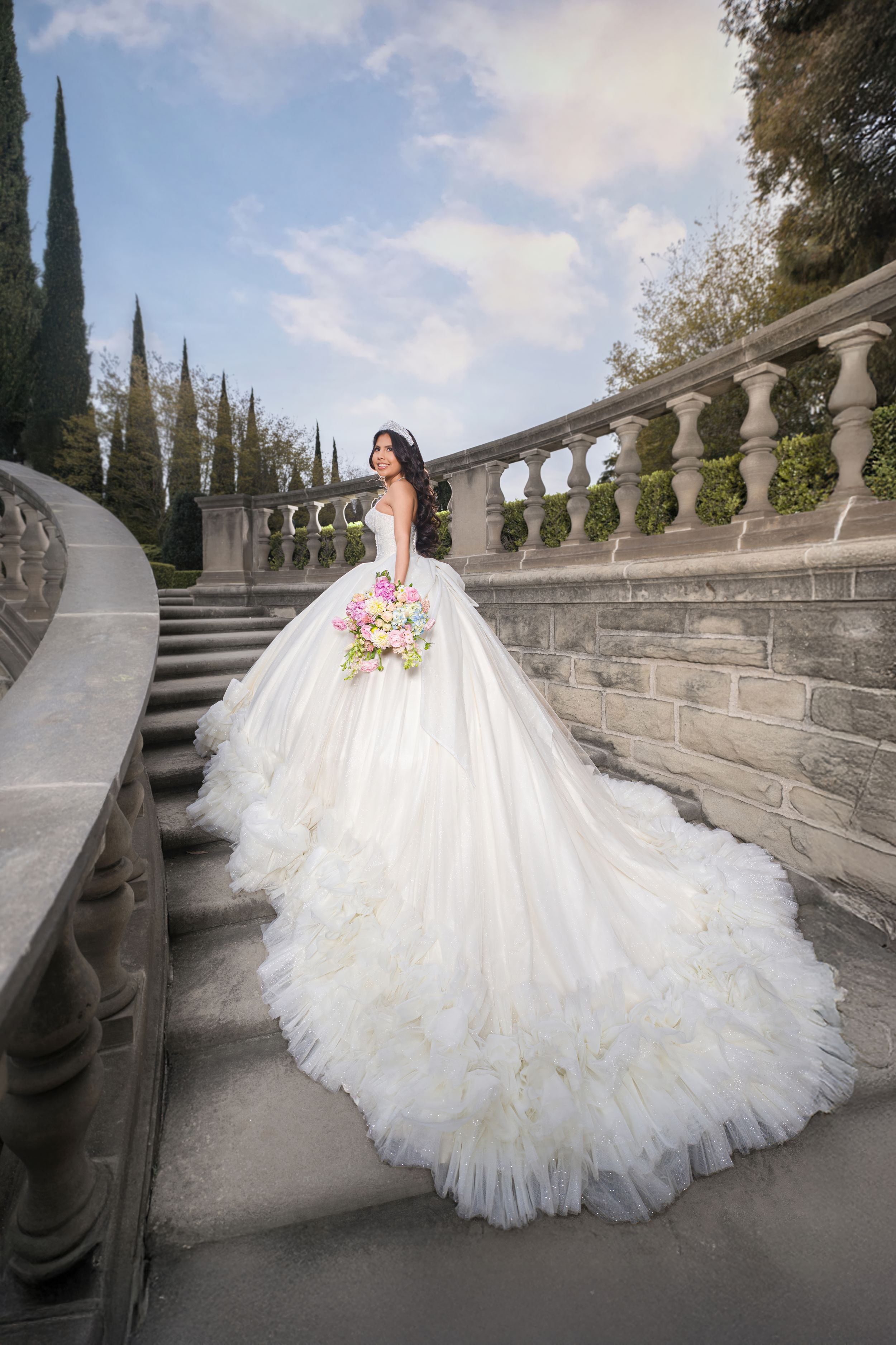 Bride in flowing white ballgown wedding dress with long train poses on elegant stone staircase lined with cypress trees.