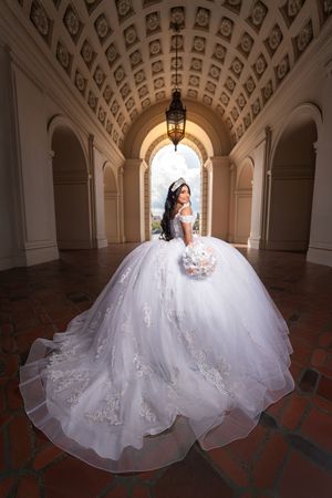 A bride in an elegant white ball gown poses in a grand arched hallway with ornate ceiling details and warm lighting.