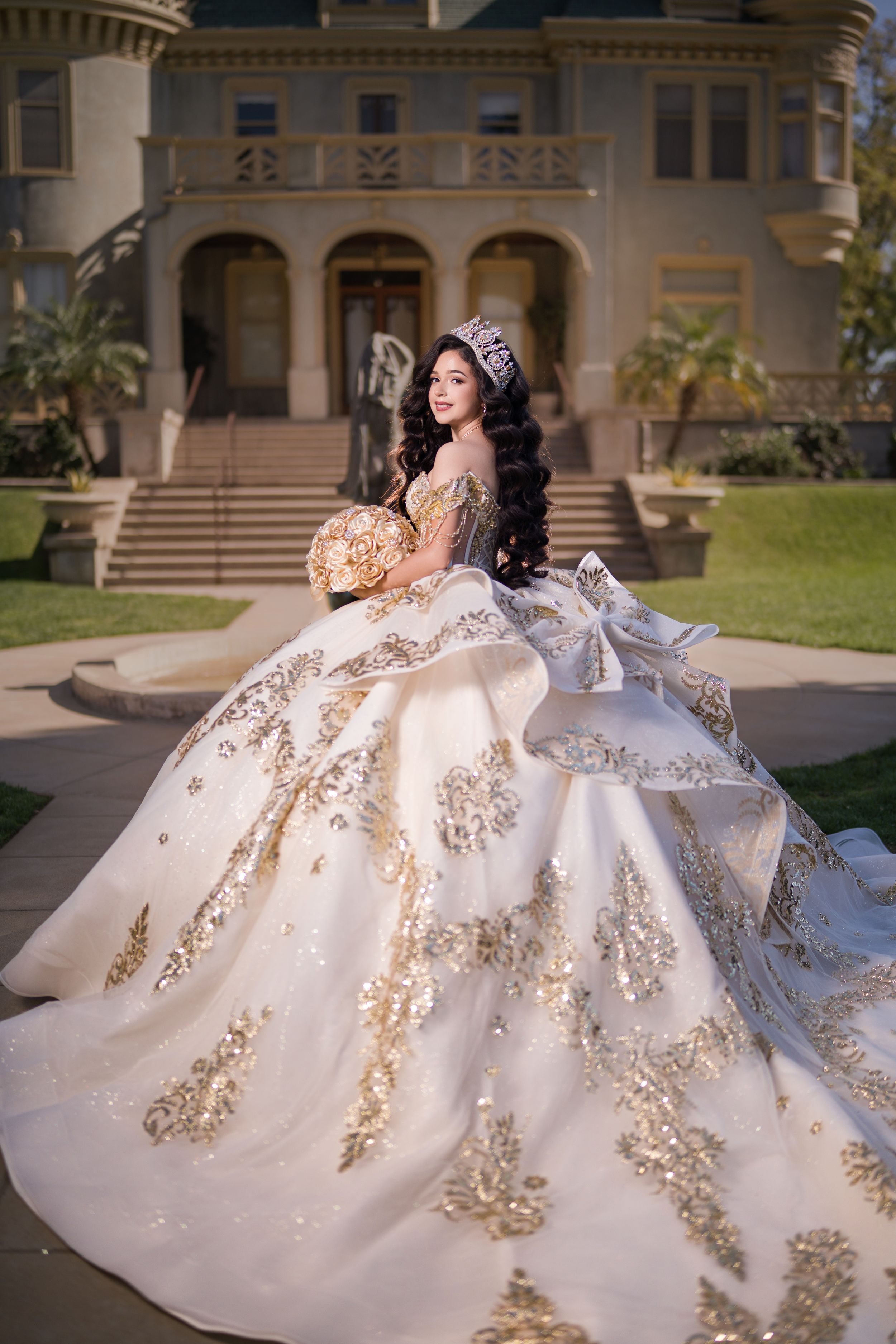 Young woman in white and gold quinceañera ball gown with tiara holds bouquet before a grand mansion.