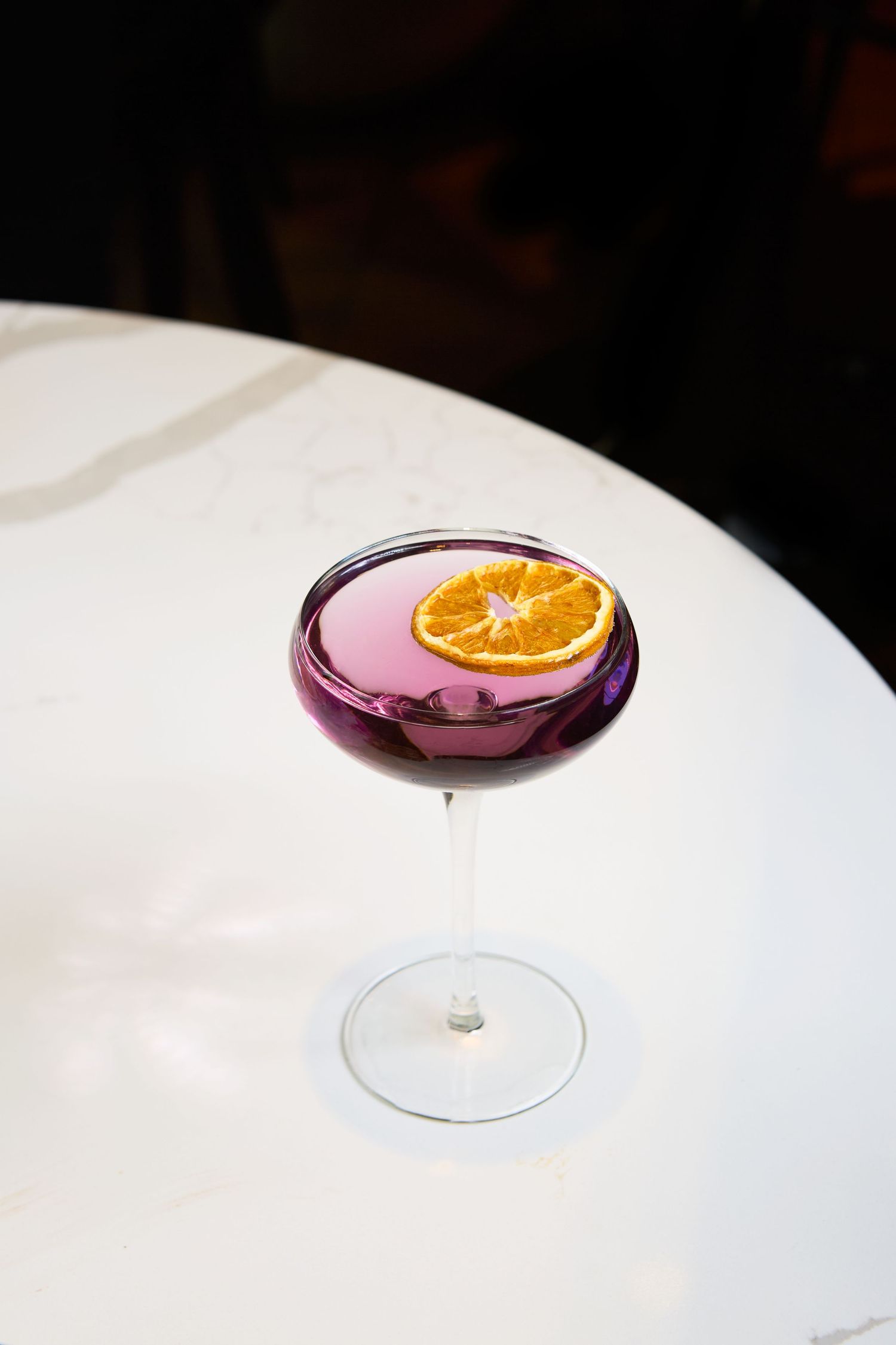 A purple cocktail in a coupe glass garnished with a dehydrated orange slice on a white table.