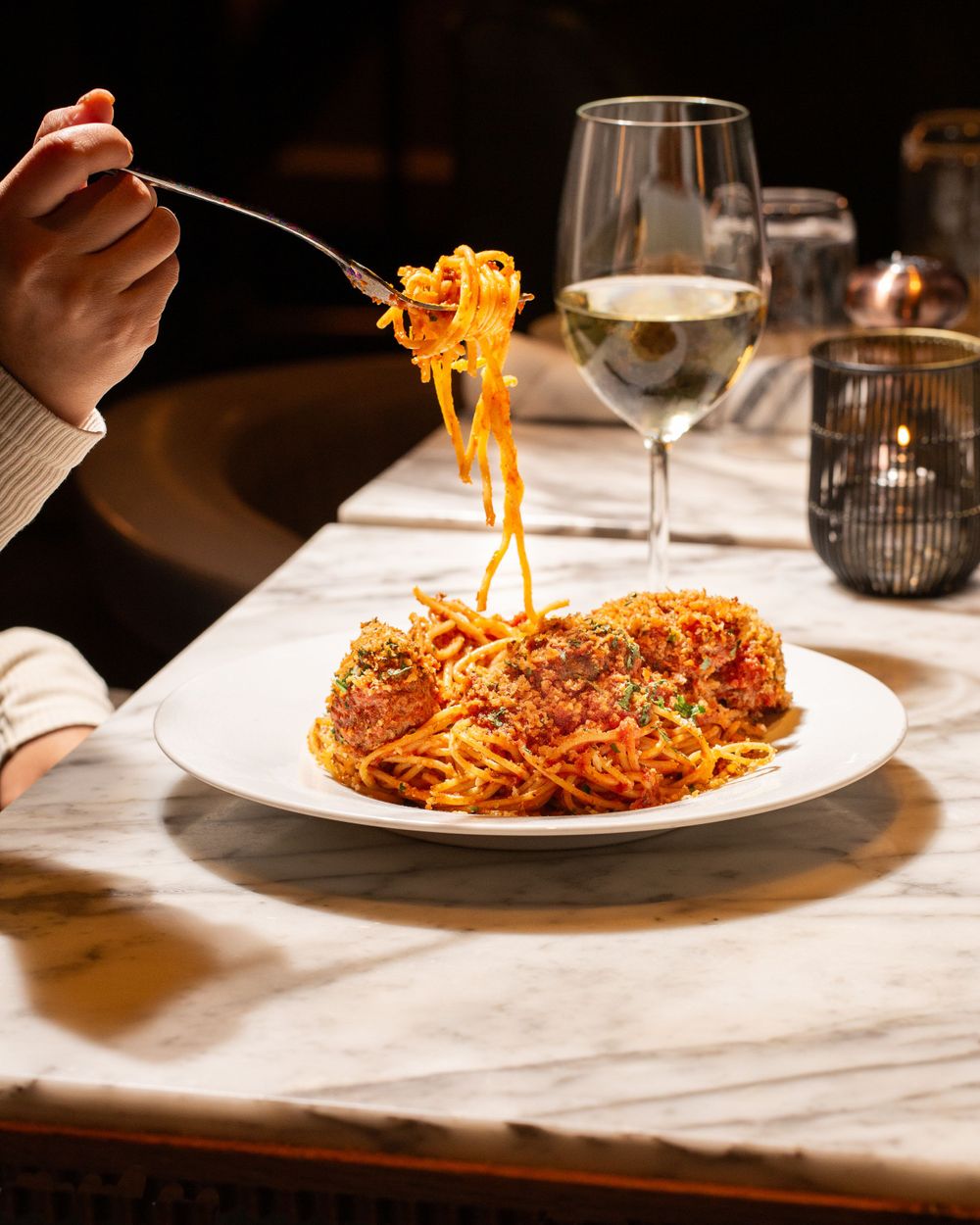 A plate of spaghetti with meat sauce being twirled on a fork, alongside a glass of white wine on a white tablecloth.