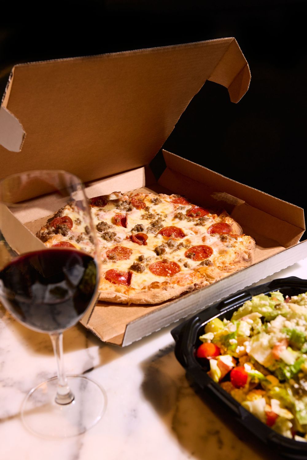 A pepperoni pizza in a delivery box served with red wine and a side salad with vegetables.