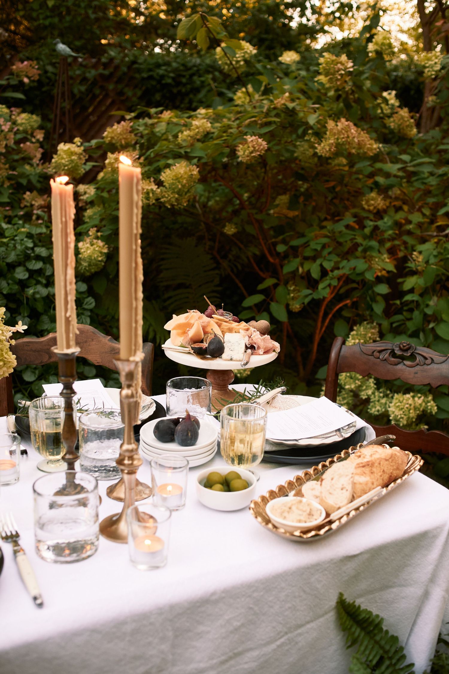 Elegant outdoor table setting with tall candles, pastries, and glassware against lush garden greenery at sunset.