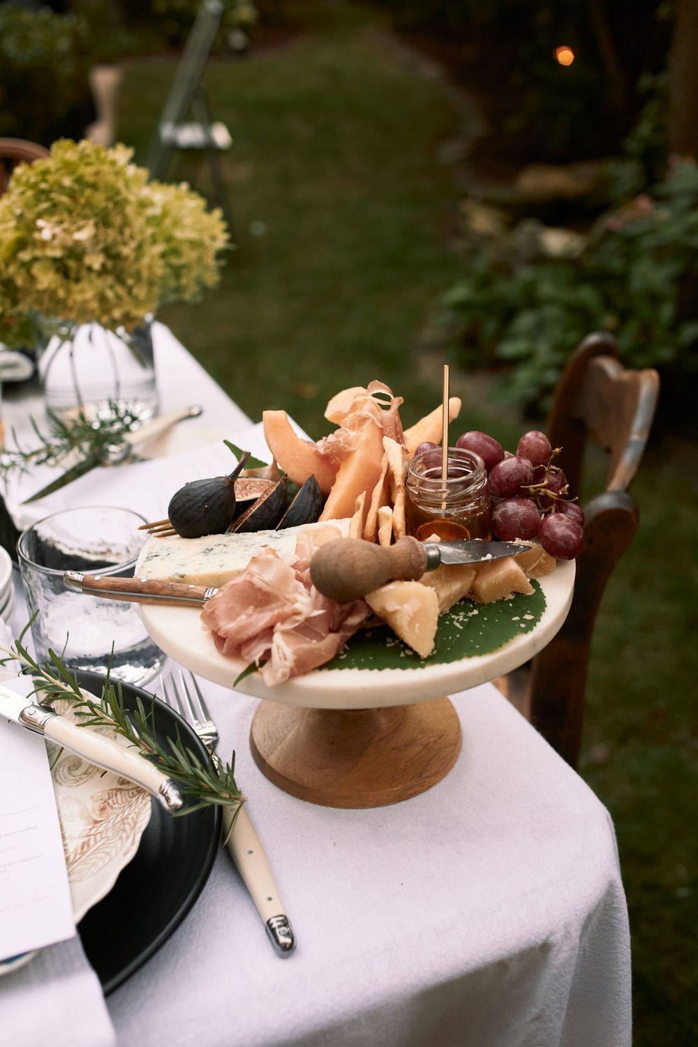 An elegant charcuterie display on a raised stand with prosciutto, grapes, and flowers on a white tablecloth outdoors.