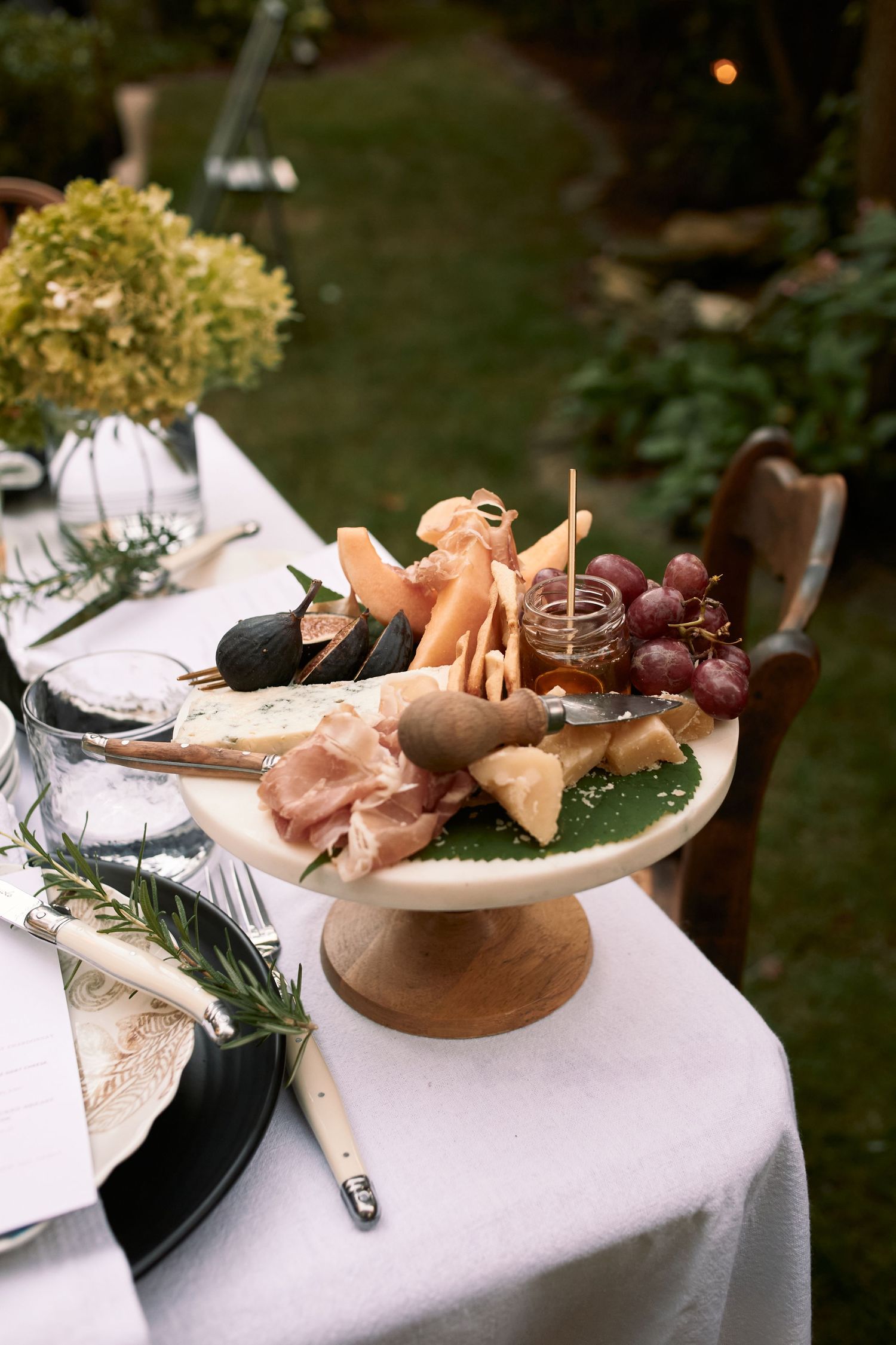 An elegant charcuterie display on a raised stand with prosciutto, grapes, and flowers on a white tablecloth outdoors.
