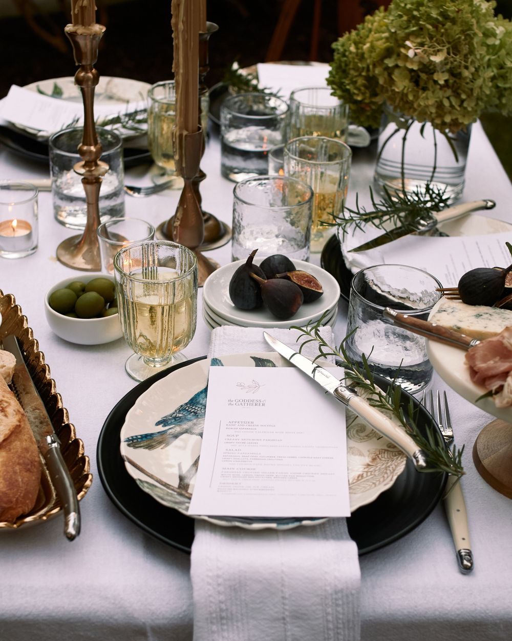 Elegant dinner table setting with candlesticks, olives, menu cards and wine glasses on white linen tablecloth.