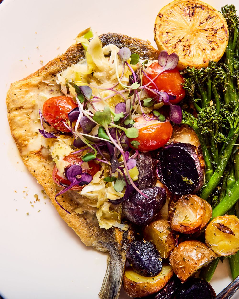 Grilled fish fillet with colorful microgreens, roasted potatoes, cherry tomatoes and charred lemon on a white plate.