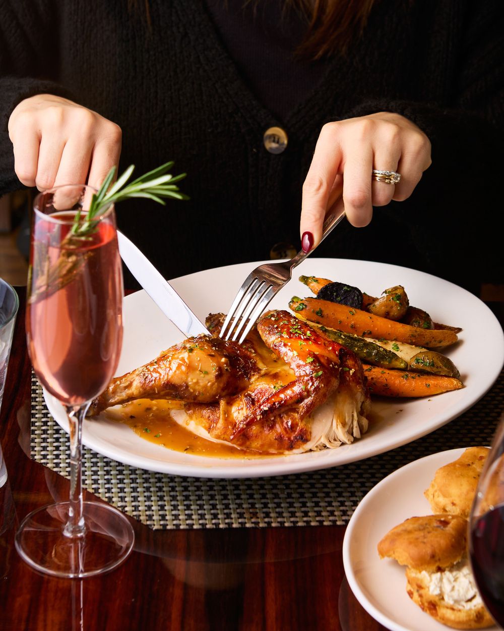 Elegant dinner plate with glazed chicken and vegetables alongside a pink cocktail garnished with rosemary on dark table.