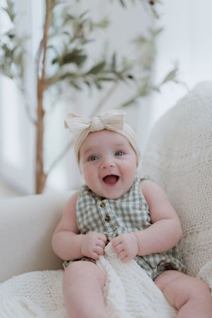 Baby wearing a gingham romper and bow headband smiles joyfully while sitting on white couch.