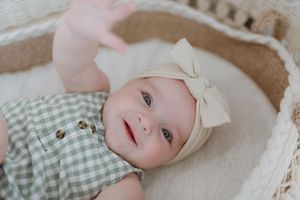 A baby wearing a cream bow headband and mint green gingham outfit lies on soft white bedding.