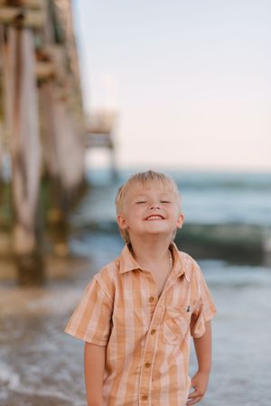 A child wearing a striped peach shirt smiles joyfully on the beach with a pier in the background.
